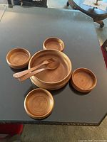 Photo of the full 6-piece wooden salad bowl set displayed on a dark table, showing one large bowl, four smaller bowls, and two wooden utensils (spoon and fork).
