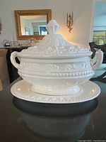 Front view of Victorian-style white ceramic tureen with lid and matching underplate placed on a table, well-lit setting.