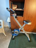 Side view of the XTERRA foldable exercise bike showing silver frame with blue accents, padded seat, pedals, and black stabilizers on carpet