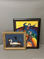 Photo showing both oil paintings together, the smaller goose decoy still life in a decorative gold frame and the larger Superman-themed painting with bright colors and dark frame.