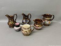 All six lusterware pitchers arranged side by side, showing various decorative styles and sizes.