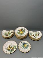 All 24 plates grouped by motif and shape arranged on a flat surface, showing detailed hand-painted fruit designs and gold trim.