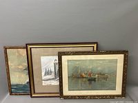 Three framed lithographs leaning against a wall depicting coastal scenes: two with boats and one with a lighthouse in winter.