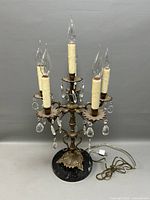 Full view of the brass candelabra table lamp with five candle-style bulbs, visible faux wax detailing, crystal prisms, and round dark base. Power cord and plug are shown.