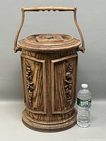Full front view of the mid-century style faux wood ice bucket with two doors decorated with carved floral details. Plastic water bottle shown for scale.