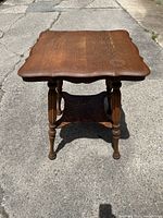 Front view of solid oak end table showing scalloped square top, turned legs, and lower shelf with carved detailing, sitting on concrete.