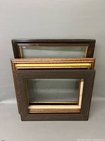 Four stacked picture frames with glass. The top frame is dark brown showing wear. Frames beneath feature gold-tone and other finishes.