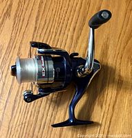 Dark blue and silver Ex Series fishing reel with fishing line on wooden surface photographed from the side showing handle.