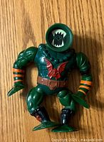 Front view of the vintage green action figure showing circular head with monster face, muscular arms, orange stripes, black boots, and bat emblem.