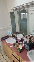 Wide shot of bathroom countertop showing sink, vanity mirror with lights, hairbrush, multiple cosmetic and bathroom items arranged on countertop.
