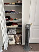 Open closet with four shelves holding folded colorful blankets, towels, fabrics, and scarves. Two large packaged blankets or linens on floor. A closed-door closet with a gray tower fan standing outside.