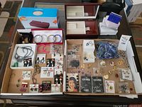 Full overview of the costume jewellery lot displayed in boxes and trays showing variety of jewelry pieces including earrings, bracelets, rings, brooches, and necklaces.