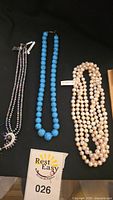 Three necklaces displayed on black fabric: two freshwater pearl necklaces, one turquoise necklace, and a third multicolored beaded necklace with crescent pendant