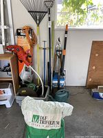 Full view of the Black & Decker GrassHog trimmer and edger along with garden tools including rake, shovel, extension cord, watering can, and garden hose in gray bins.
