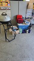 Photo showing full lot including the George Foreman grill on its stand, two gray storage cabinets, large blue Igloo cooler, red insulated bags, boxes of dishes and other camping supplies.