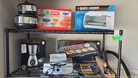 Wide shot of kitchen shelf displaying boxed Black & Decker toaster oven, Pyrex storage containers, Paderno blender, a box with griddle grill image, and various other kitchen appliances and accessories including mixing bowls and smaller boxed items.