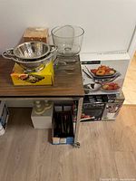 Photo showing stainless steel colanders, glass trifle bowl, and boxed cookware items on a wooden shelf