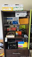 Shelf with multiple office supplies including paper reams, toner cartridge, drum unit, envelopes, folders, stamp pads, and other small office supplies
