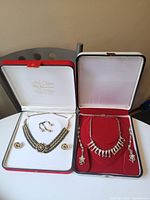 Two jewelry sets displayed side by side in open cases — one with black and gold-tone stones and flower designs, the other silver-tone with rhinestone flowers.