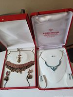 Two open red jewelry boxes each containing a necklace and earrings jewelry set; left set gold-tone with pink stones and right set silver-tone with blue stones and a ring.