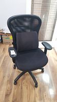Front angle view of black rolling office chair with mesh back and lumbar support cushion on wooden floor near window.