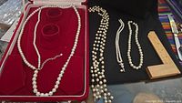 Four pearl necklaces laid out on black surface and displayed in red velvet case with two ring holders. Wooden ruler for scale included.