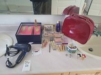 Overview photo of lot with hair dryer, red cosmetic case, multiple makeup and perfume items on white countertop.
