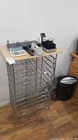 Metal standing document rack with wire racks and office items on top including a calculator, tape dispenser, and electronic devices.