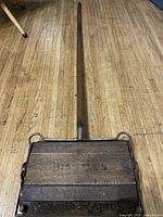Full view of the antique wooden sweeper showing the long handle and rectangular wooden body placed on a wooden floor.