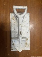 Cast-iron door knocker shaped like a shovel on a rectangular mounting plate, displayed on a wooden surface.