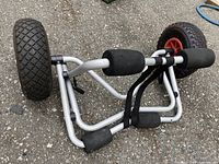 Metal boat/kayak dolly carrier laid on ground showing foam pads and large air-filled tires with red rims
