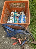 Top-down view of an orange plastic crate containing multiple tubes of construction sealants and a coiled pair of booster cables with clamps on top.