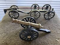 Photo showing the cast brass and silver plated miniature cannons on black wheeled carriages placed on a surface in front of window blinds
