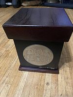 Rectangular wooden keepsake box with dark wood lid and black main body with circular metallic/native art plaque on front. Signed by Clarence D Wells 2012.