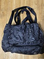 Front angled view of black shoulder bag with intricate white floral and geometric patterns, showing front zipper pocket and shoulder straps.