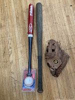 Photo showing new Rawlings red/black T-ball bat and ball in packaging next to used wooden bat and used baseball glove on wood floor