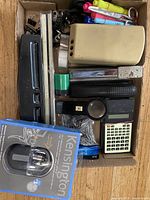 Photo of a box with various small office supplies including staplers, scissors, pens, a pencil sharpener, and a 3-hole punch.