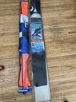 Two packaged kites side by side on wood flooring, showing orange, blue, black, and white design.