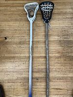 Full view of two lacrosse sticks standing side by side on wooden floor, one white with blue gradient and one wood-toned with black head.