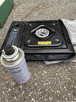 Black single-burner Sterno butane stove with visible burner and control knob, placed on ground next to one fuel canister.