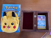 Yellow Pikachu plush purse, Pokémon Sinnoh Handbook, wooden box open with Magic cards inside on a wooden surface