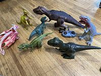 Several dinosaur toys displayed on a wooden floor including a large brown T-Rex with wired remote, and six other various dinosaur toys.