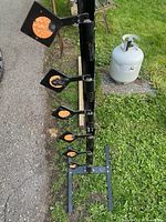 Photo of the black steel plate target stand with six swinging orange targets arranged vertically on a grass area, propane tank visible for scale.
