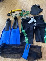 Photo shows swimming gear comprising two blue swim fins, black and green Speedo swim jammer, two pairs of goggles, snorkel, and mesh bag arranged on a wood floor.