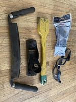 Photo showing all four items: vintage draw knife, block plane, yellow scraper, and new safety glasses in packaging laid on wooden surface.
