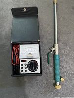 Vintage analog multi tester with visible dial, test leads stored inside black carrying case next to hand valve.