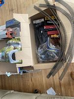 Box with curved and straight train tracks, model station building, painted landscape tunnel, and plastic bag with accessories.
