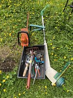 Photo of orange gas-powered weedeater, white and green electric hedge trimmer and a black tub containing various garden hand tools placed on grass with yellow flowers.