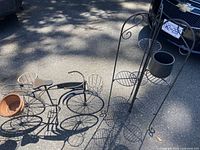 Two wrought iron plant stands shown outdoors on asphalt with shadows; bike-shaped stand includes terra cotta pot and multi-level stand is tall with circular wire shelves and decorative scrollwork.