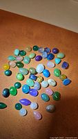 Photo showing a variety of polished natural gemstones arranged on brown surface in mixed colors including green, blue, purple, pink, yellow, and brown. Stones are cabochon cut in oval, teardrop, and round shapes.